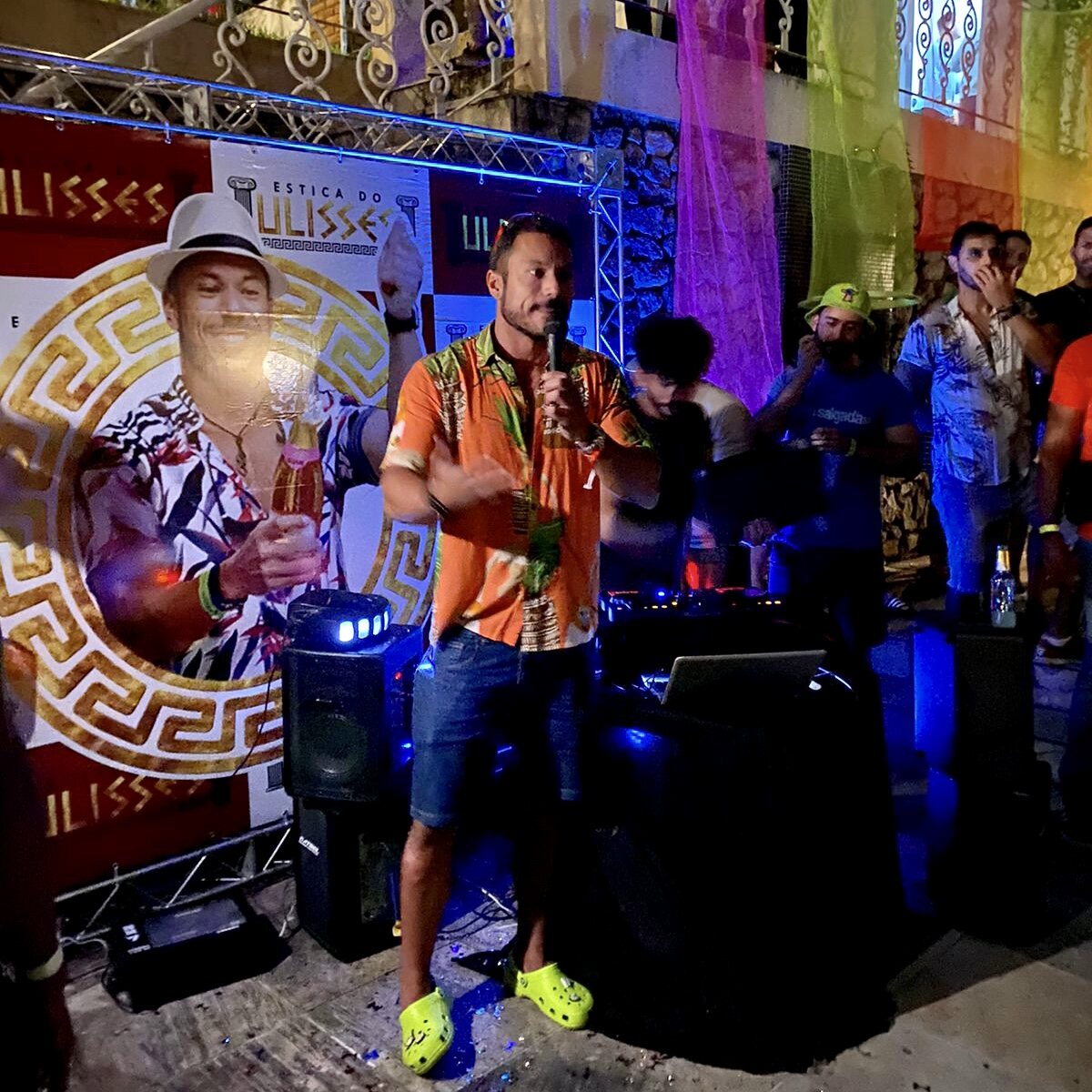 Ulisses no microfone no palco iluminado com banner Estica do Ulisses ao fundo